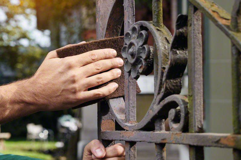 A person is sanding a rusty metal gate railing with sandpaper. Late summer daylight in the background.