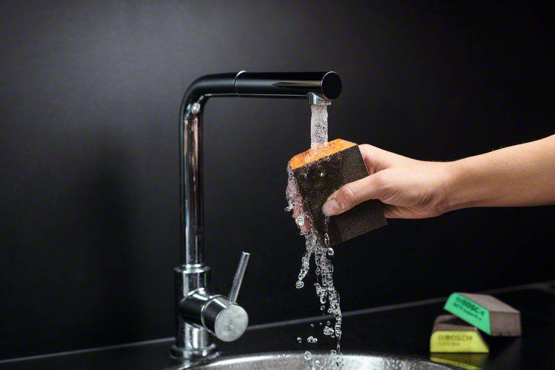 A hand holds a sponge under running water from a modern tap in a kitchen.