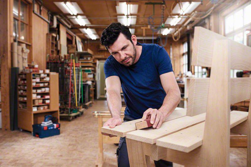 A man is working intently in a workshop, sanding a wooden board. Tools and shelves can be seen in the background.