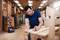 A man is working intently in a workshop, sanding a wooden board. Tools and shelves can be seen in the background.