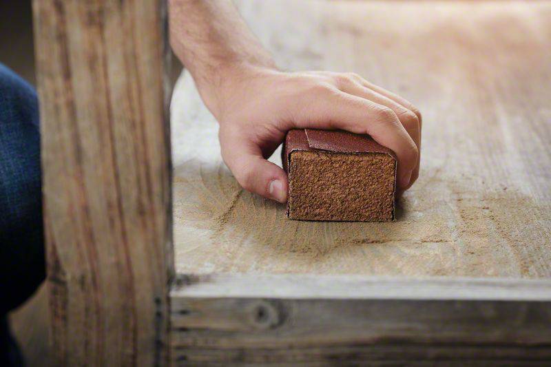 A hand holds a sanding block and works on the surface of a piece of wood. The movement follows the direction of the wood grain.
