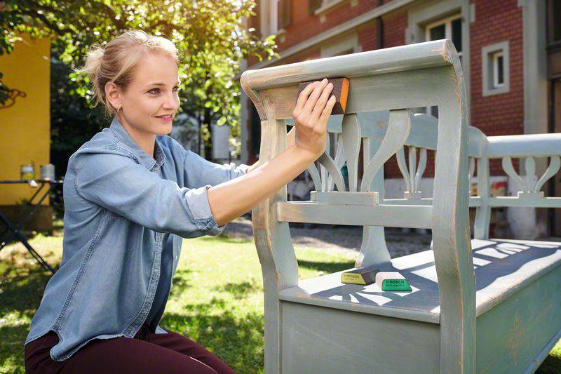 A woman is sanding a blue wooden bench in the garden. She is concentrating on her work. Trees and a house are visible in the background.
