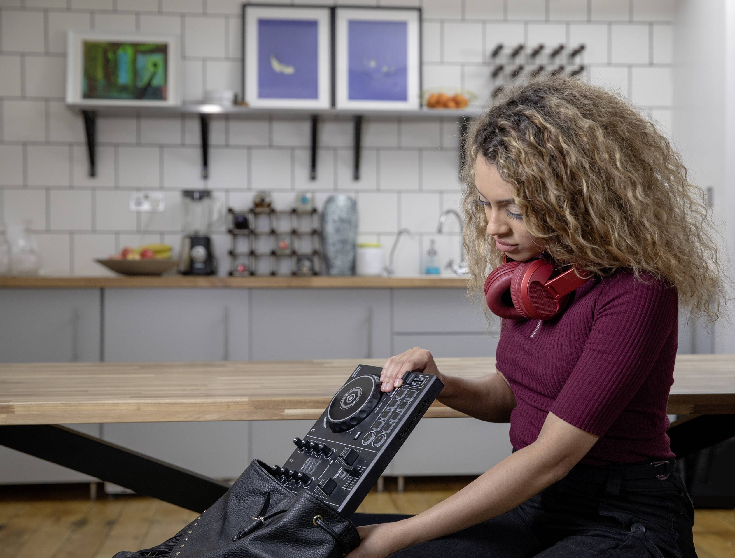A woman is sitting at the table in a modern kitchen, wearing headphones around her neck and packing a DJ mixing desk into her rucksack.