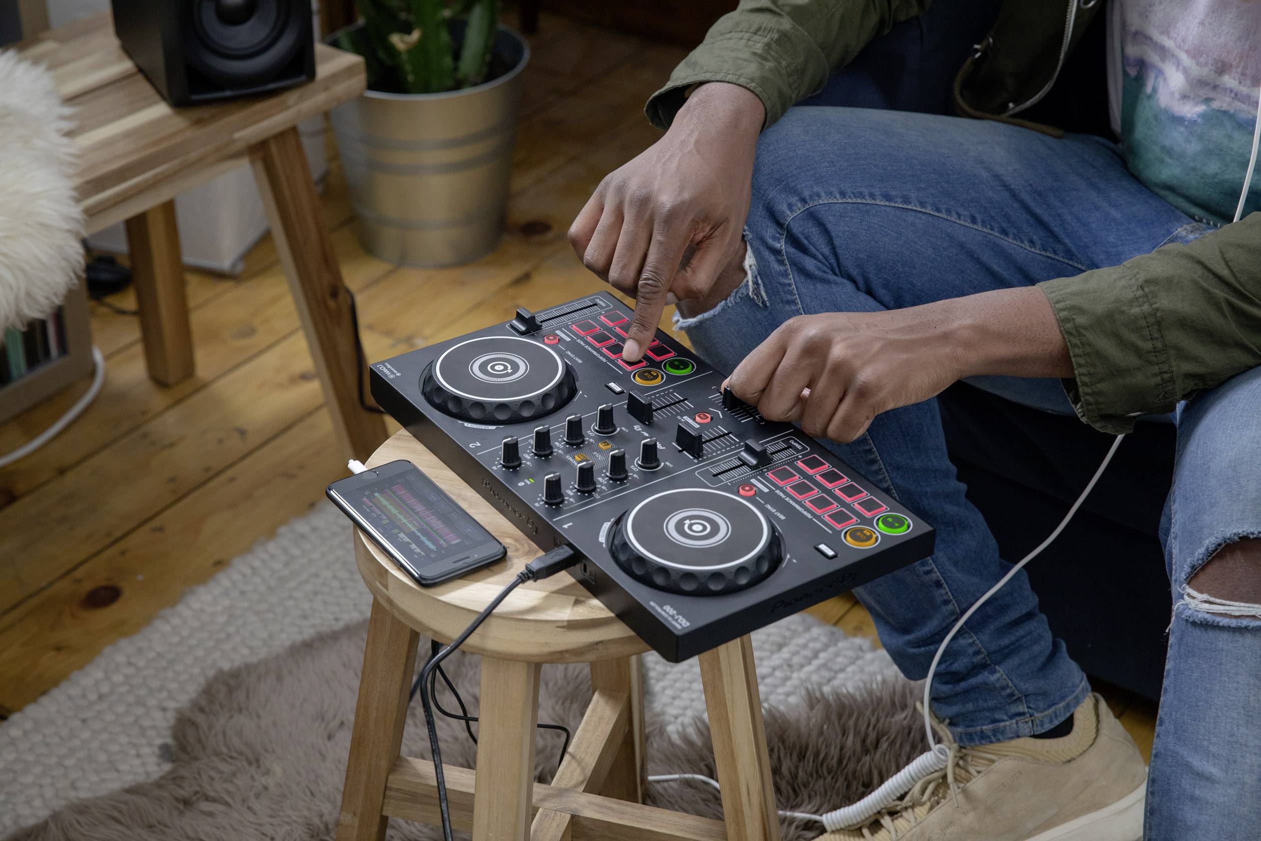 A person is operating a DJ mixing desk on a small wooden table. A smartphone is lying on the table. The room is cosily furnished.