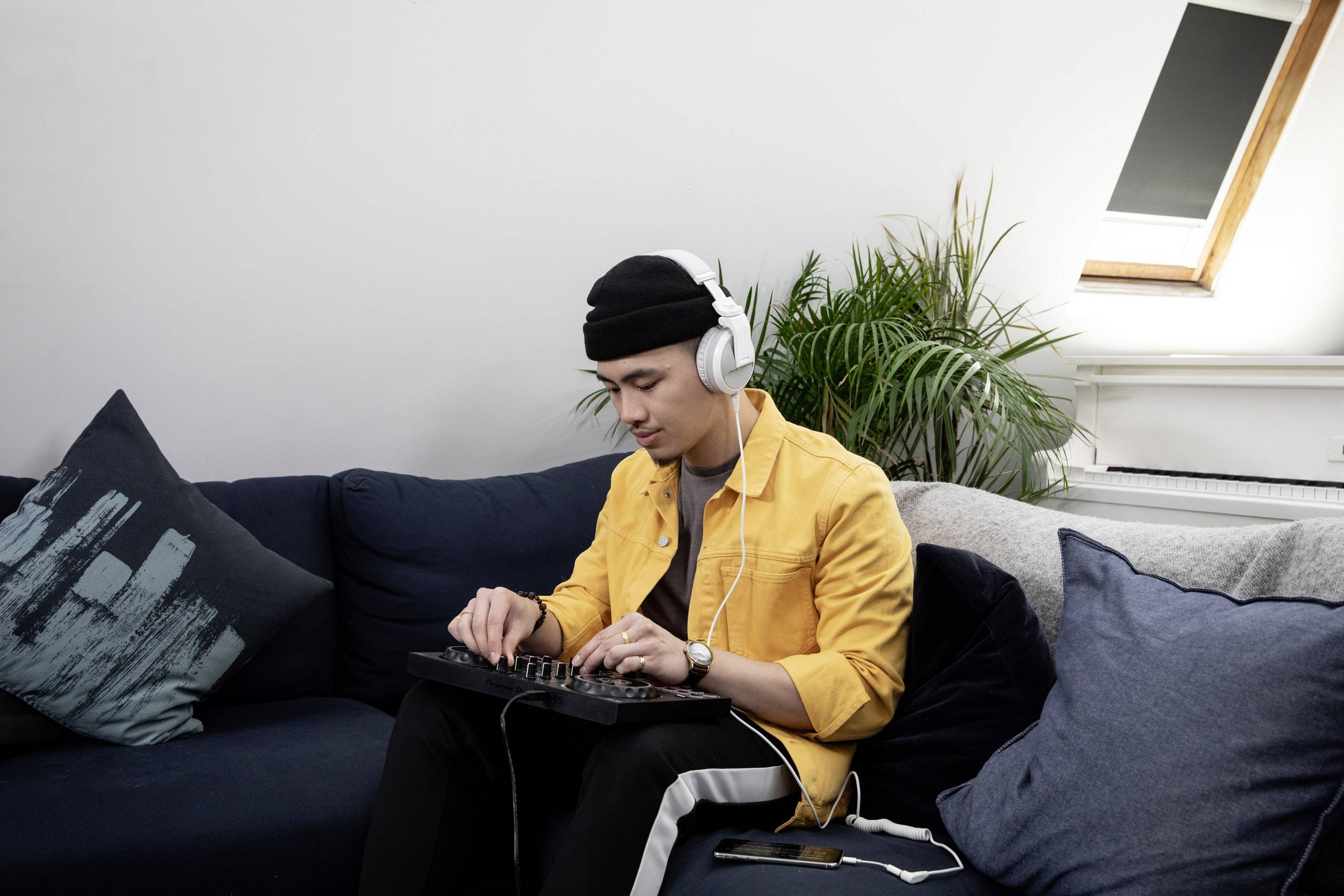 A young man sits on a sofa, wearing headphones and working intently on a mixing desk. Cushions are lying beside him.