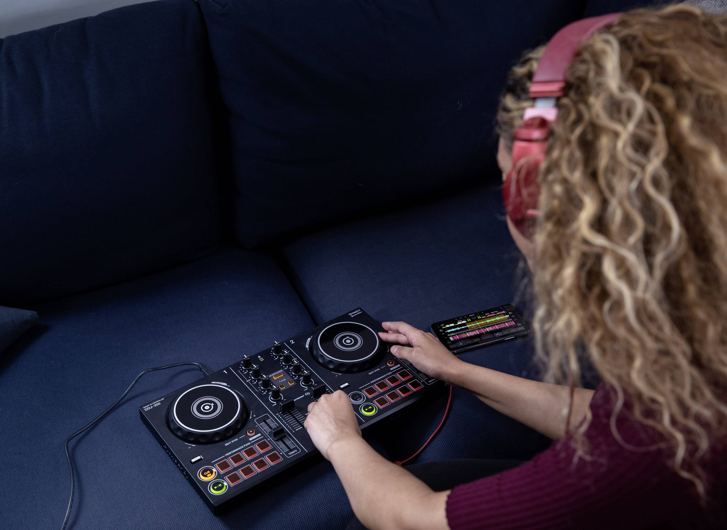 A person wearing headphones is mixing music on a DJ controller while sitting on a sofa, operating various buttons and controls.