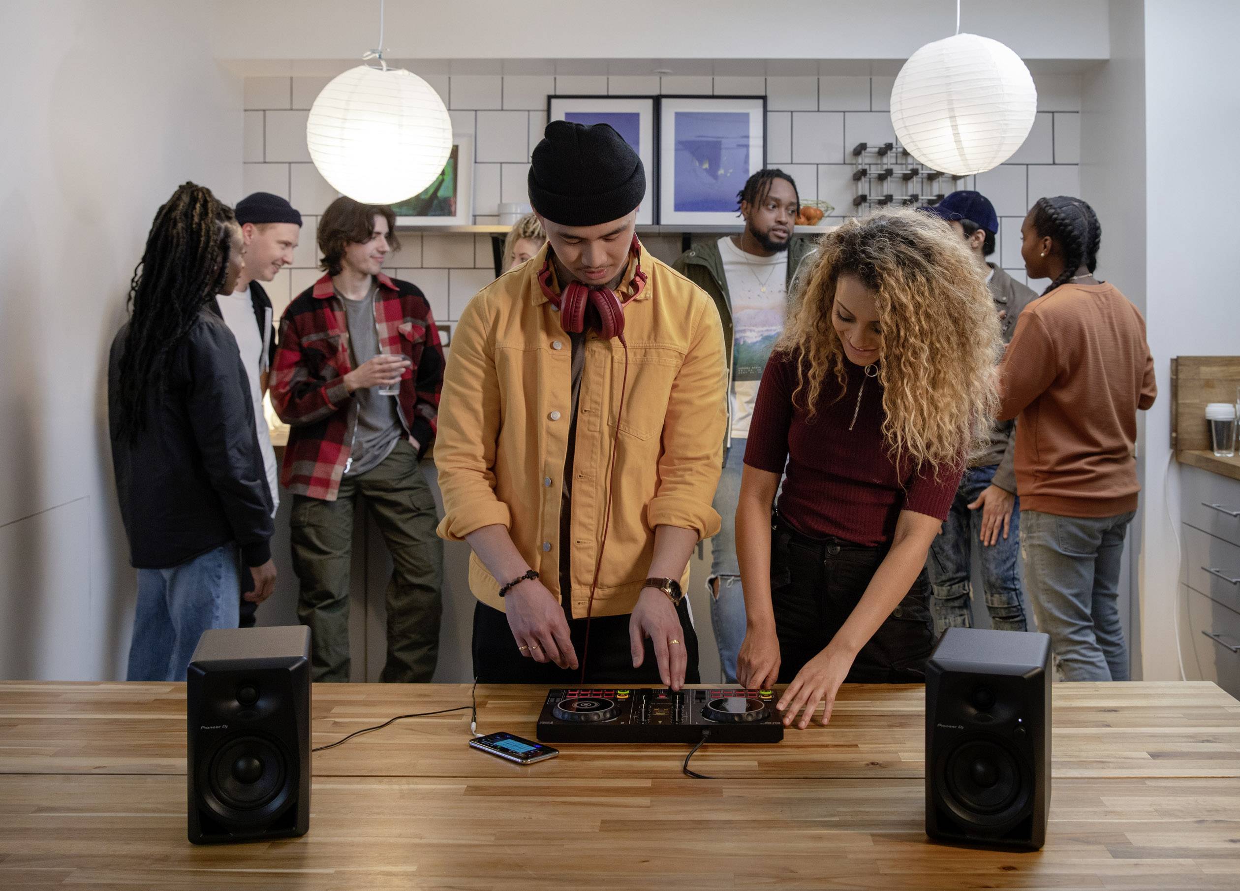 A group of people in a kitchen, two individuals are mixing music at a DJ deck, while others are chatting and drinking.