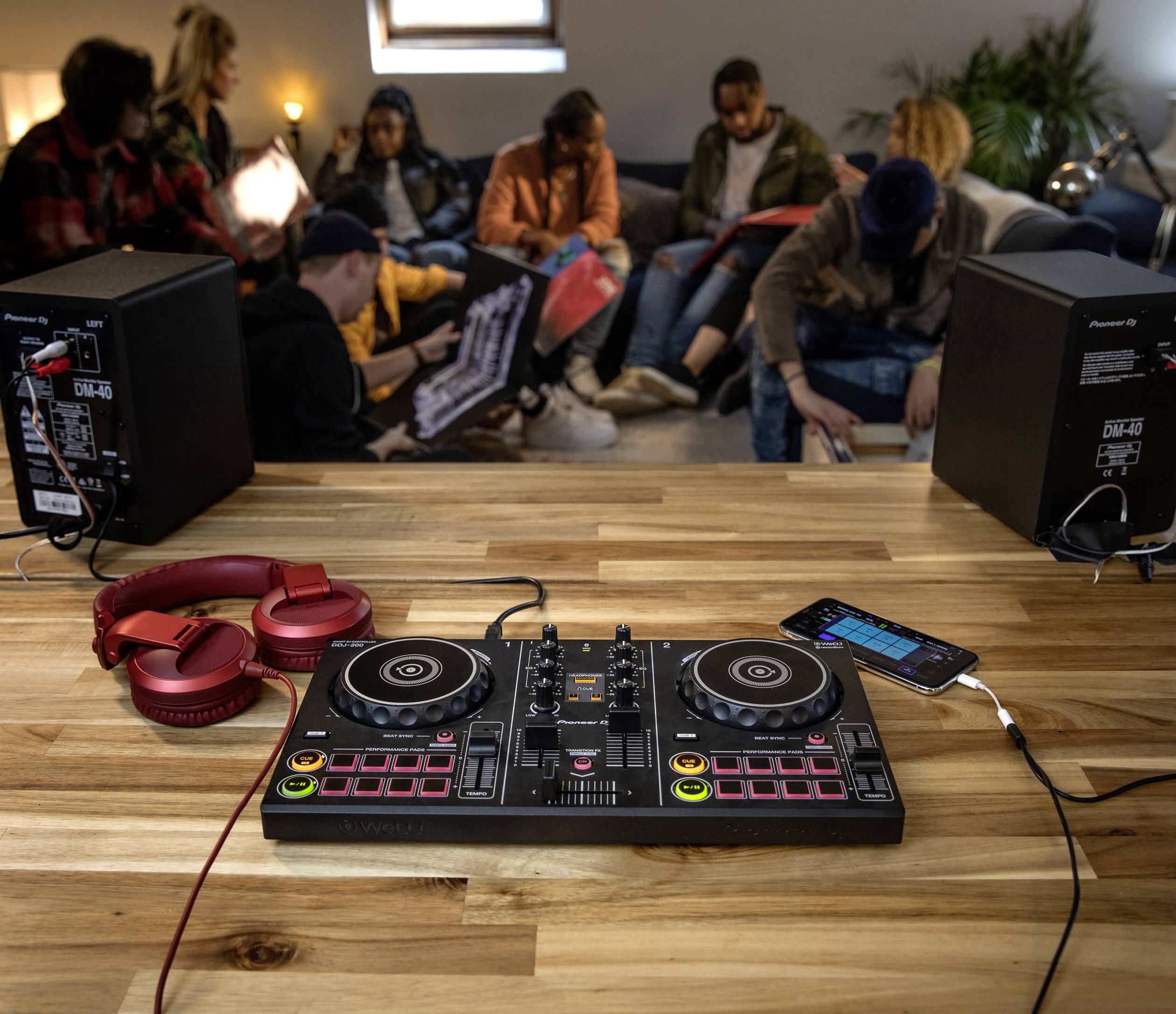 DJ controller on a wooden table with headphones and smartphone. In the background, several people are sitting on sofas and chatting.