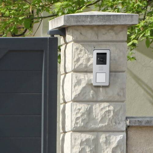 A stone gatepost beside a gate with an intercom system attached, set against a green background.