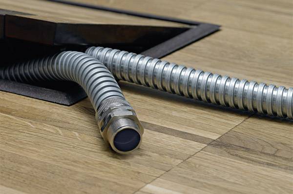 A metal cable conduit emerges from an open floor hatch in a wooden floor.