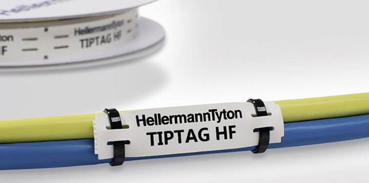 A white cable label with the inscription 'HellermannTyton TIPTAG HF' is attached to yellow and blue cables. A spool is visible in the background.