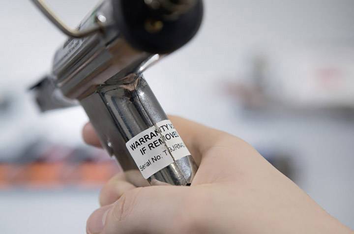 A hand is holding a device with a label that reads: 'WARRANTY VOID IF REMOVED'. The device appears to have a serial number.