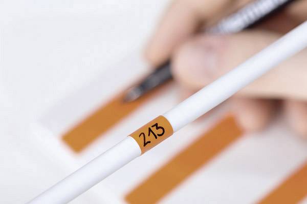 White measuring probe with the inscription '213' in the foreground. In the background, a hand is writing with a pen on a paper with orange stripes.