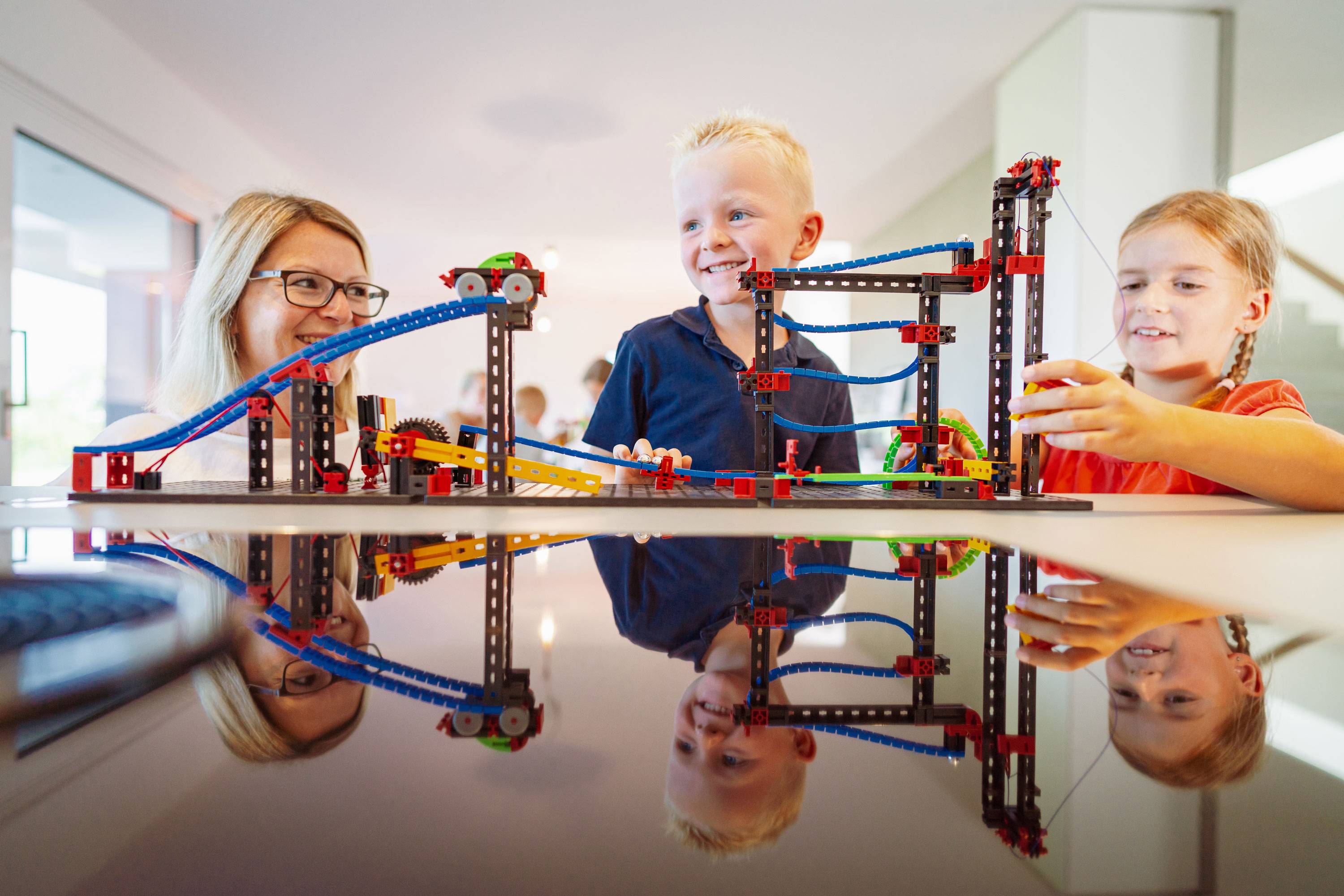 Three children are playing happily with a colourful Lego rollercoaster on a table. They are laughing and showing their enjoyment of playing together.