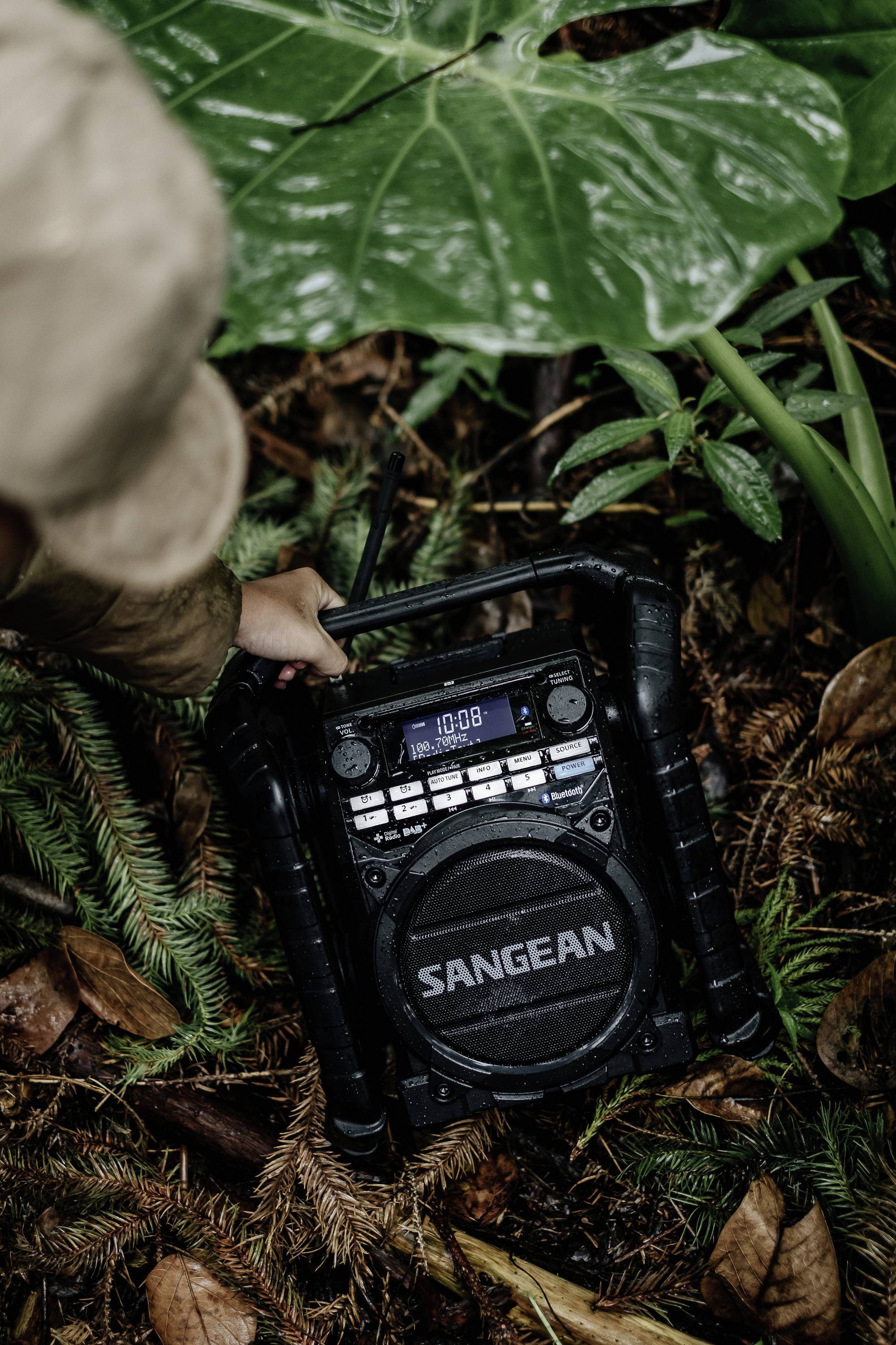 A person is holding a portable rugged radio speaker in the jungle. The radio displays '10:08'. A player is positioned on the forest floor.