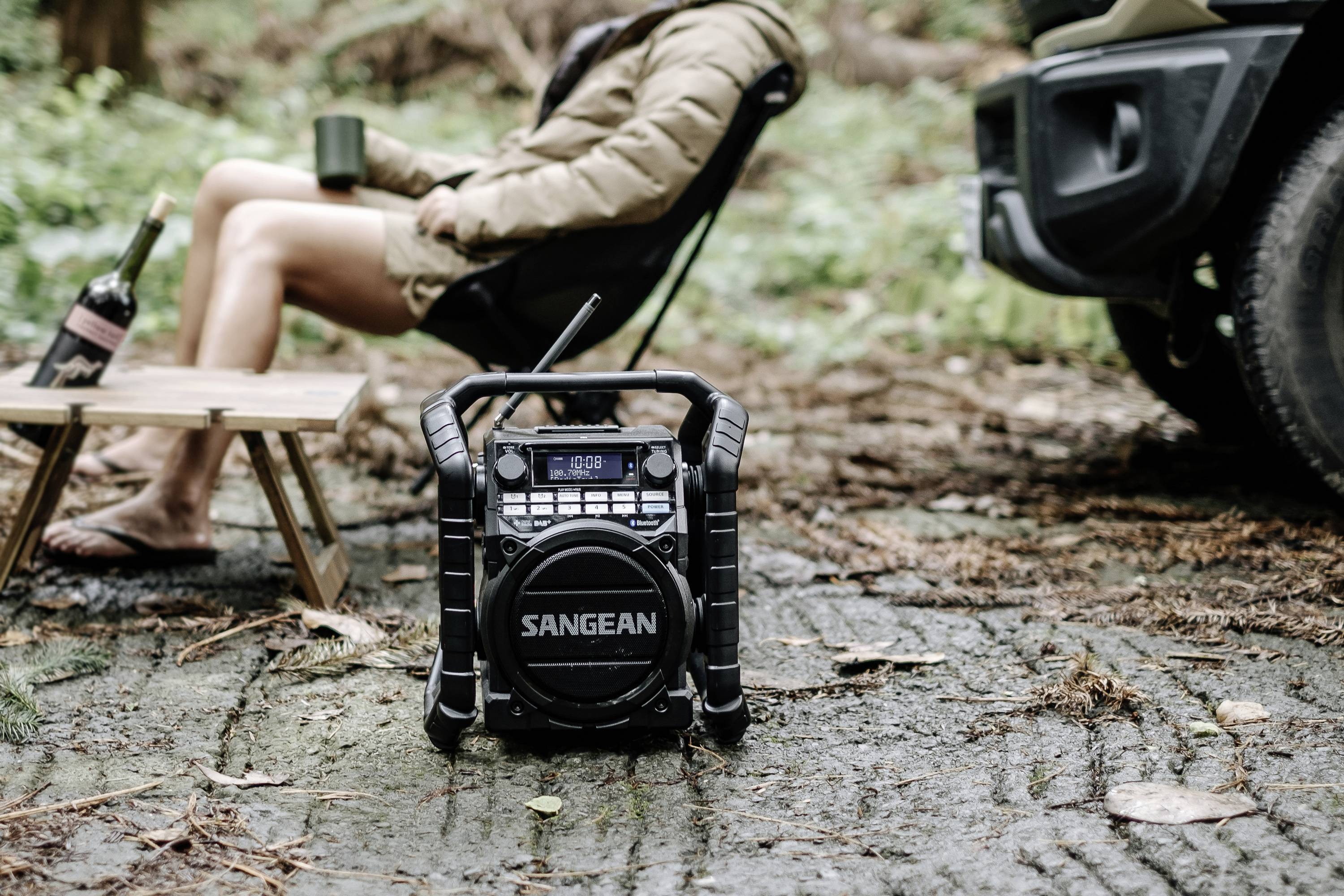 A person sits relaxed on a camping chair in the woods, holding a mug. A radio is positioned in the foreground on the ground.
