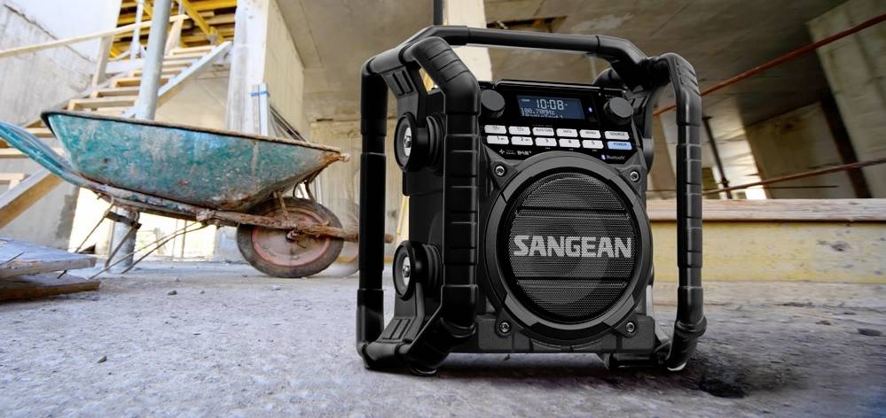 Portable construction site radio on a building site floor, next to a wheelbarrow. Robust and functional for music or news reception.