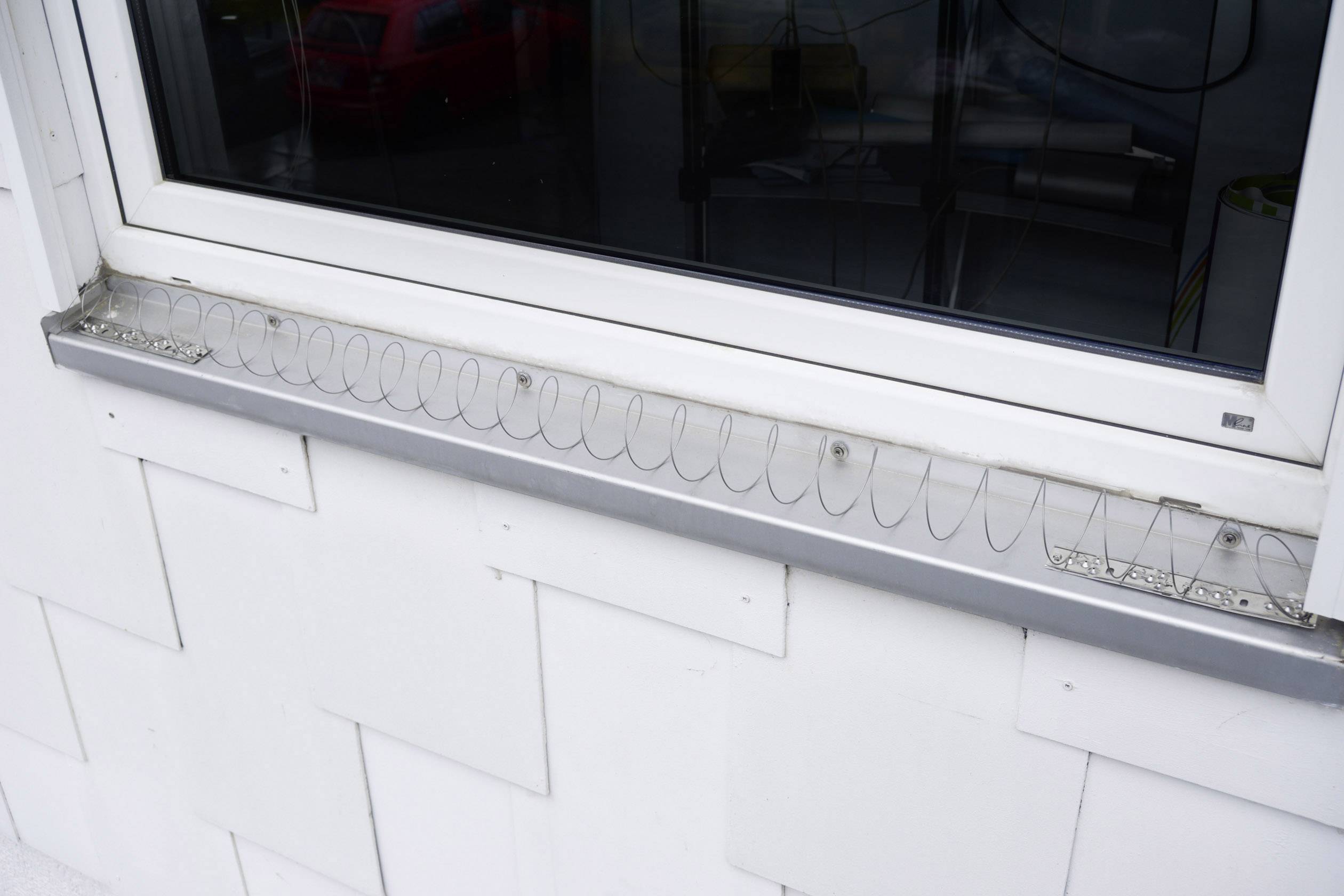 Window with an anti-pigeon wire system on the windowsill to deter birds. Cars can be seen in the background.