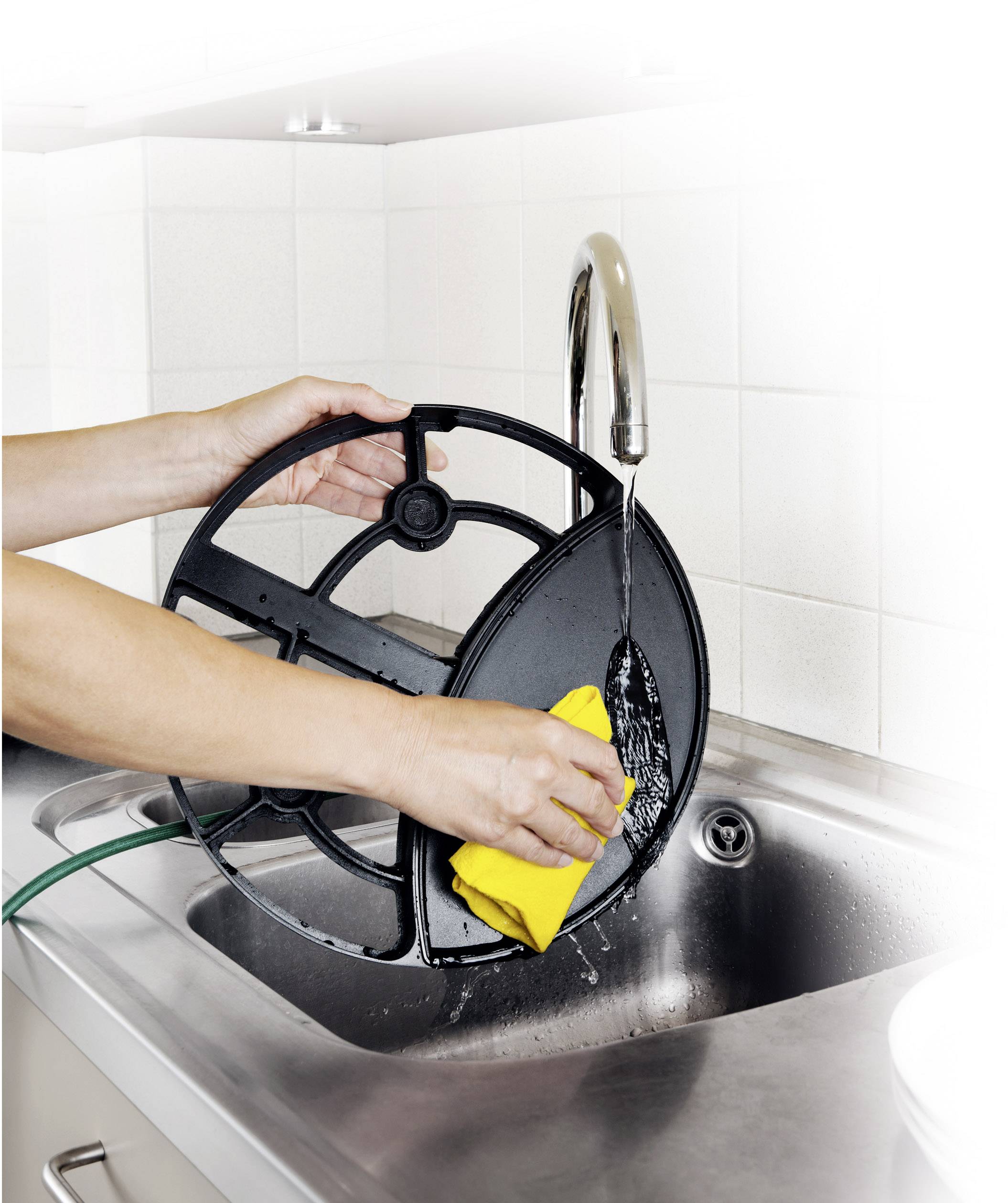 A person is cleaning a large, black kitchen appliance above the sink with a yellow sponge and running water.