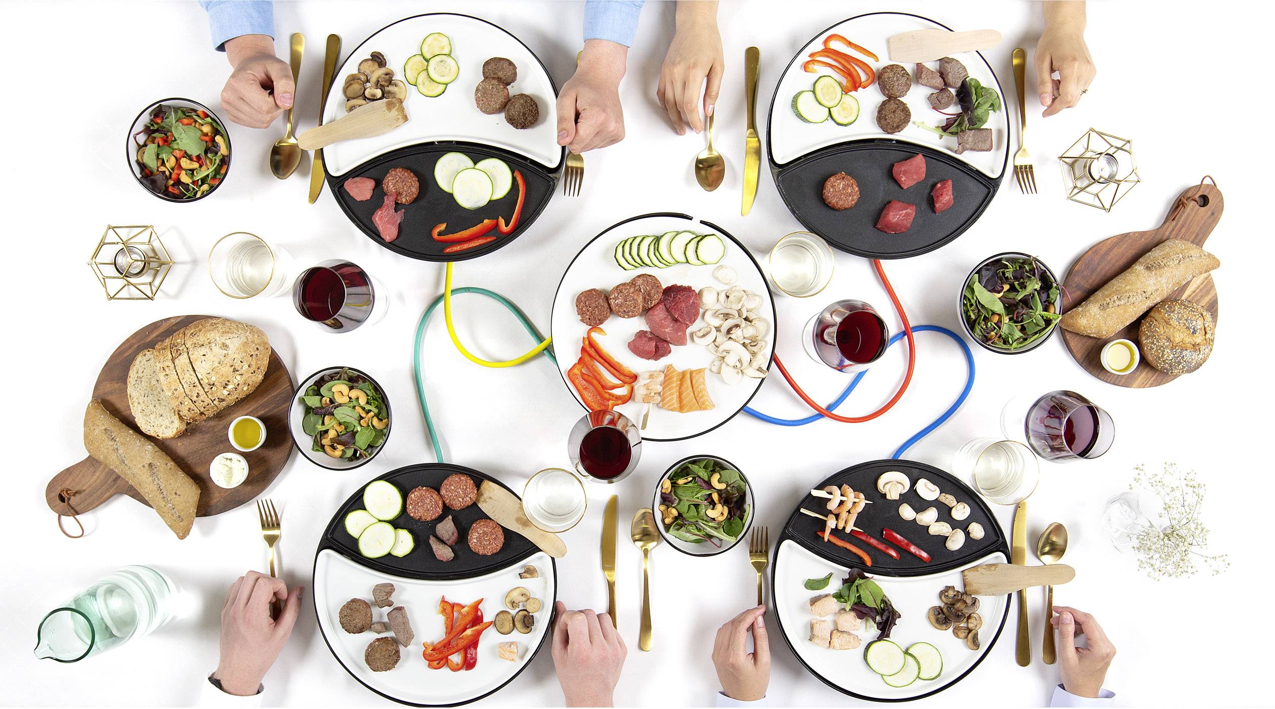 Four plates with various ingredients such as meat, vegetables, and side dishes, each plate arranged differently. Hands reach for cutlery.