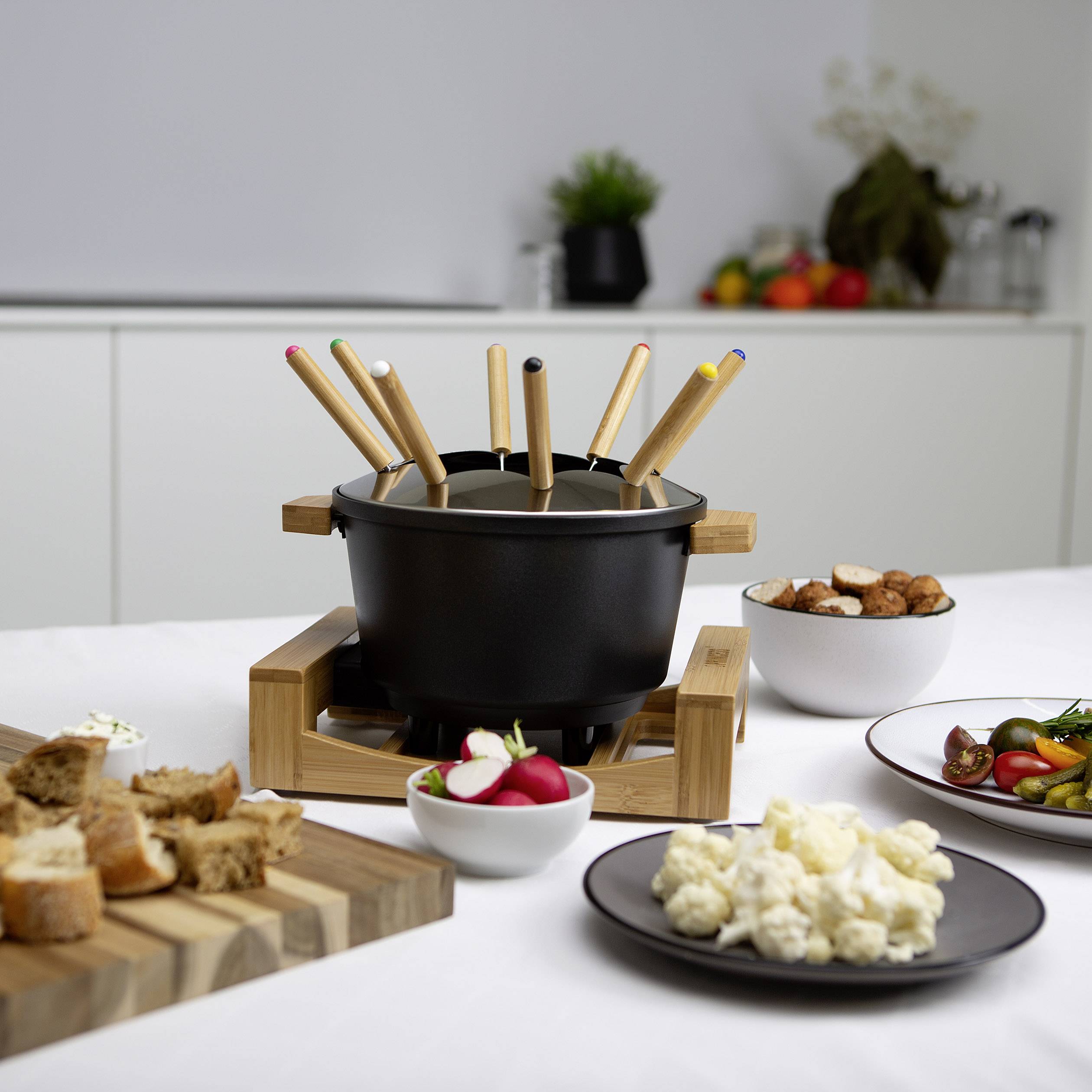 A fondue set with pots and wooden skewers on a table. In the background, plates with vegetables, bread and sauces can be seen.