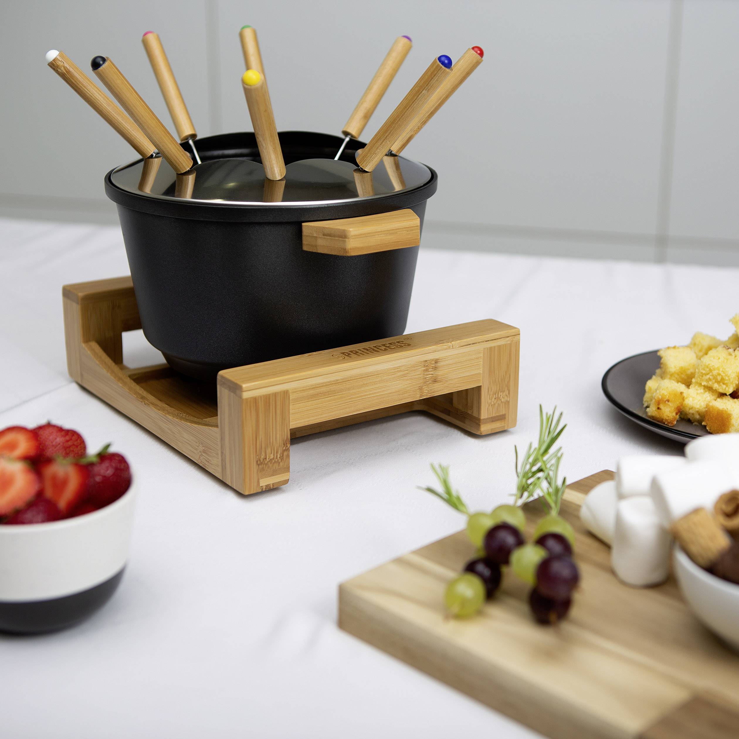 A fondue pot with several fondue forks sits on a table. Beside it are fruit bowls and small snacks, ready for a fondue meal.