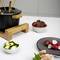 A fondue set with a wooden stand sits on a table. Beside it are bowls with courgette pieces, beetroot, and a black plate with radishes.