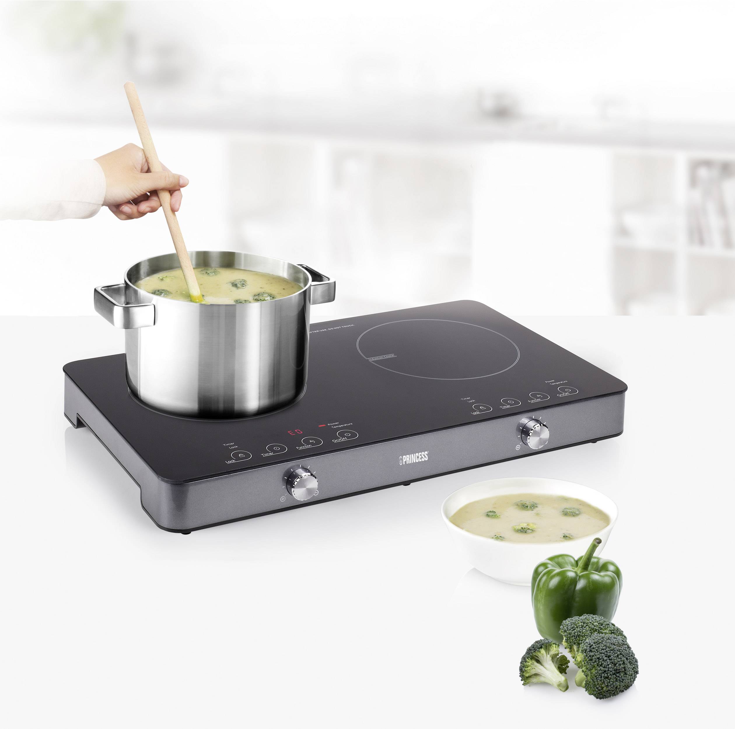 A hand stirs the soup with a wooden spoon in a pan on a modern induction hob. Next to it are peppers and broccoli.