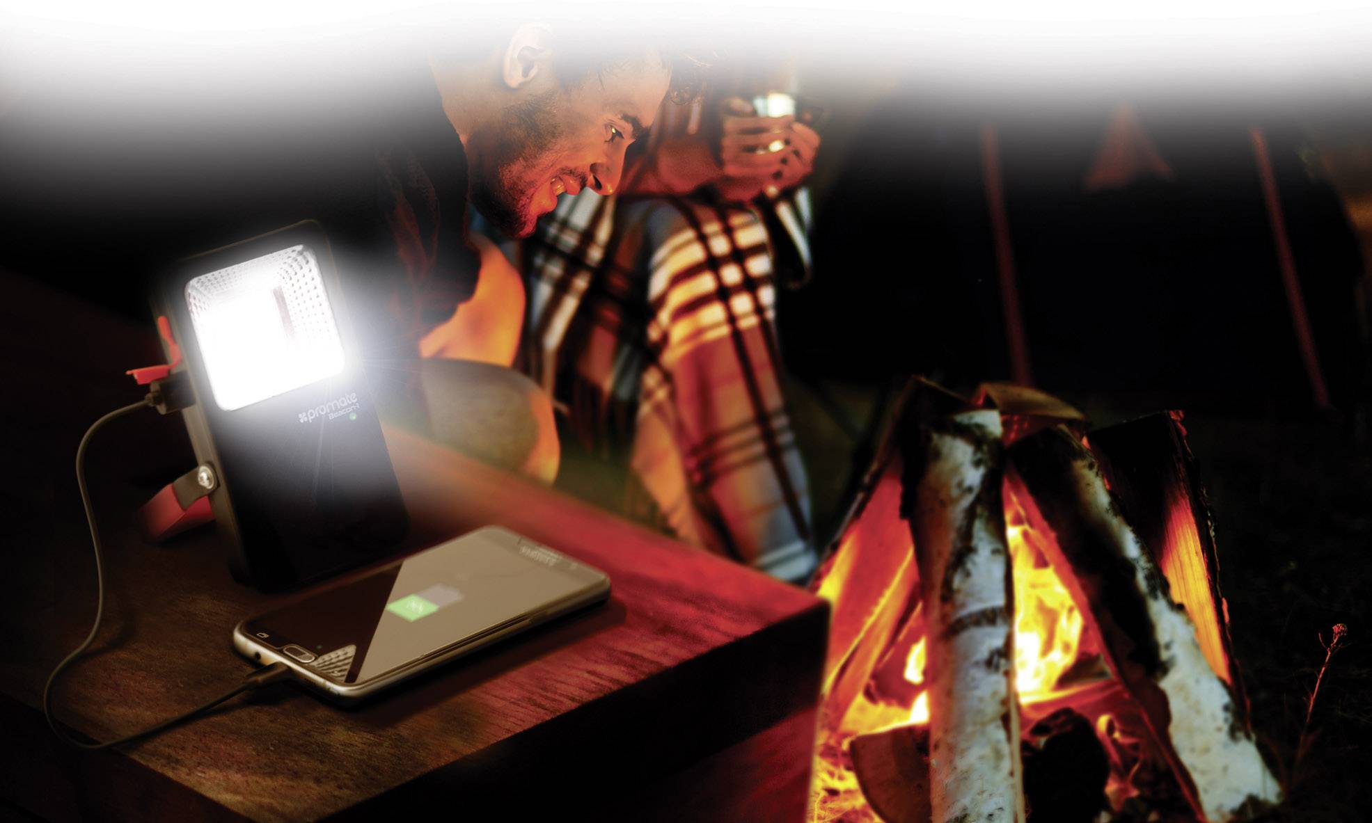 A man and a woman are sitting by a campfire. A smartphone is being charged with a portable charger on a table.