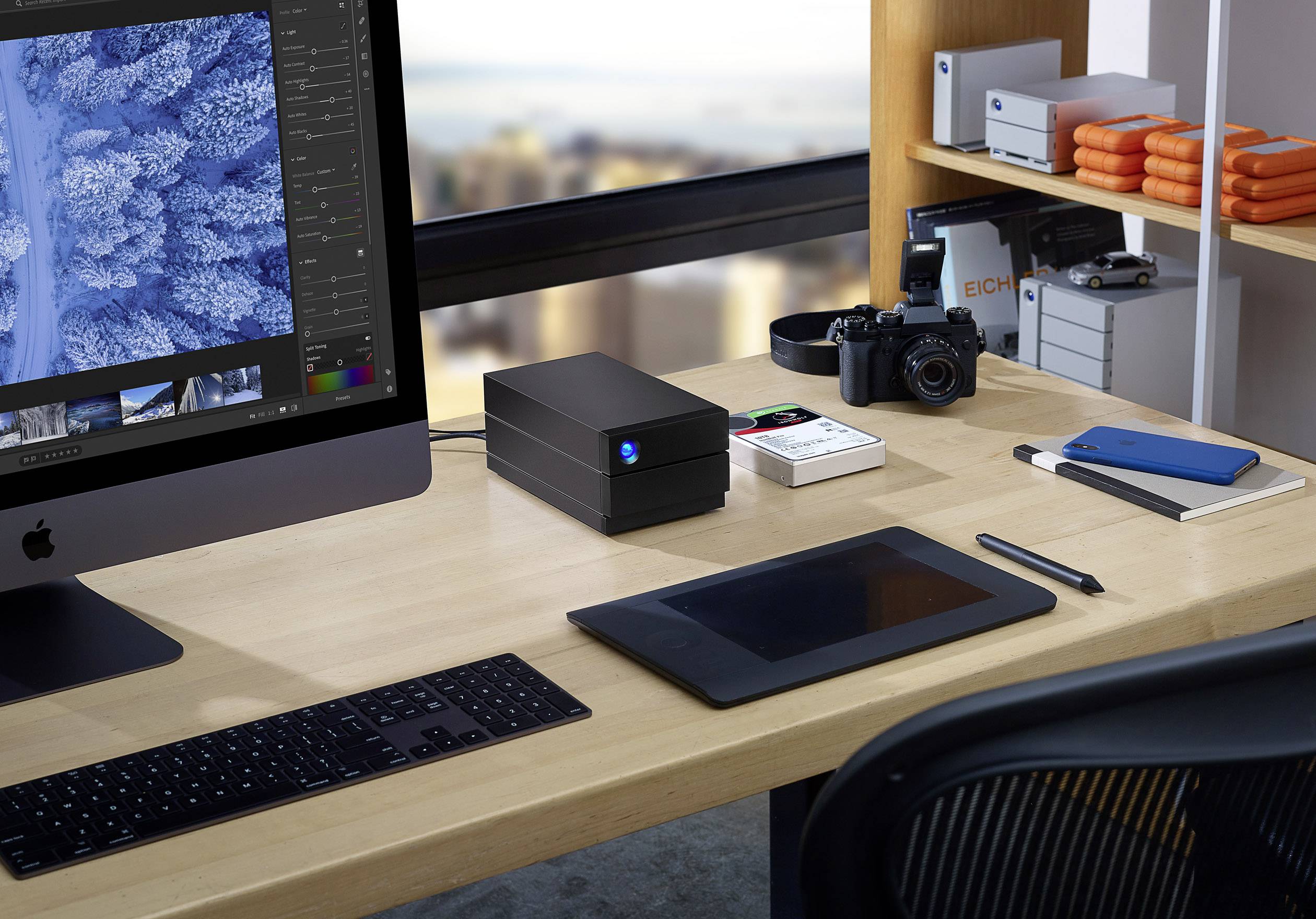 'Office workspace with a computer, graphics tablet, camera and hard drives on a desk in front of a large window with a city view.'
