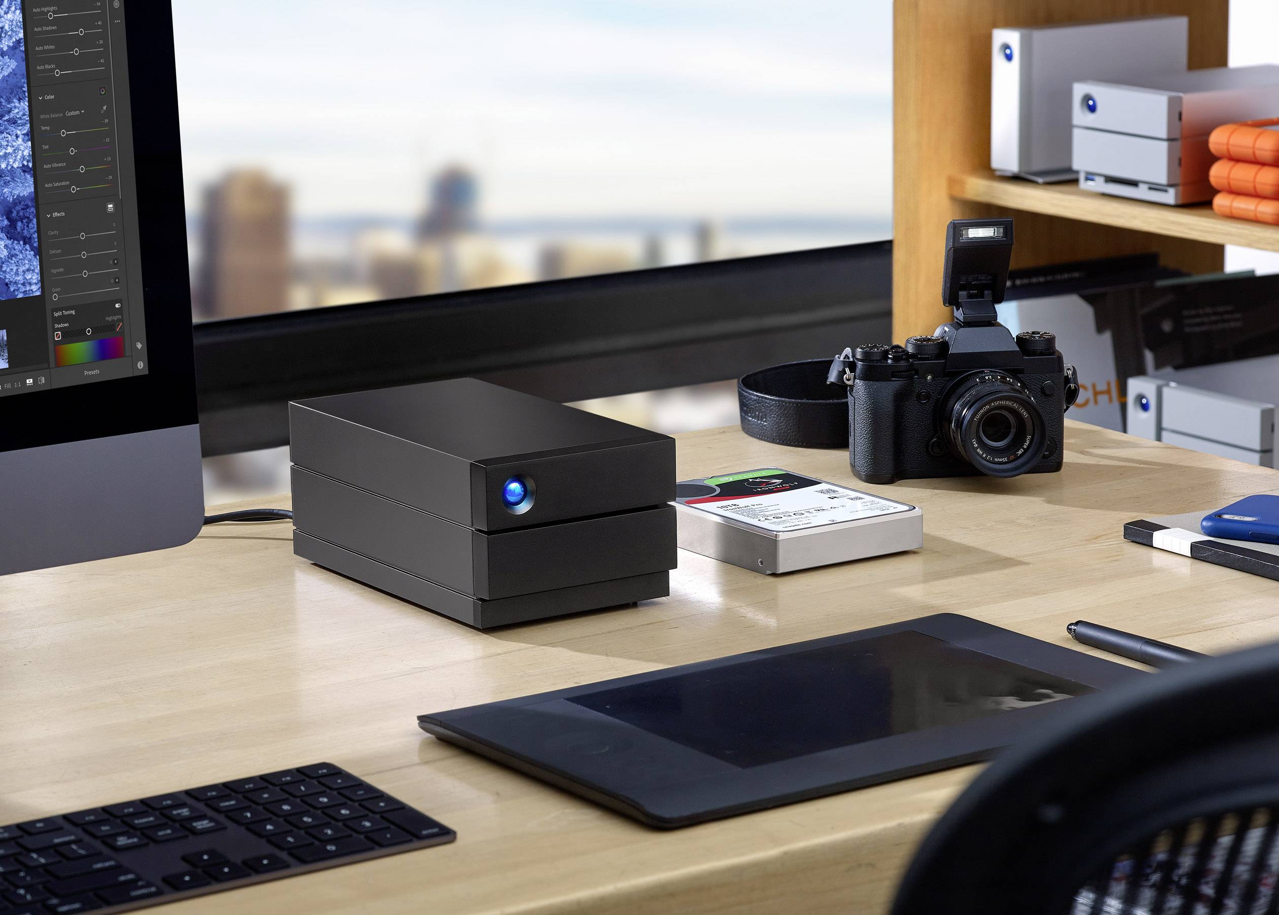 'Photo of a modern desk with an iMac, external hard drive, graphics tablet and camera, positioned in front of a window with a city view.'