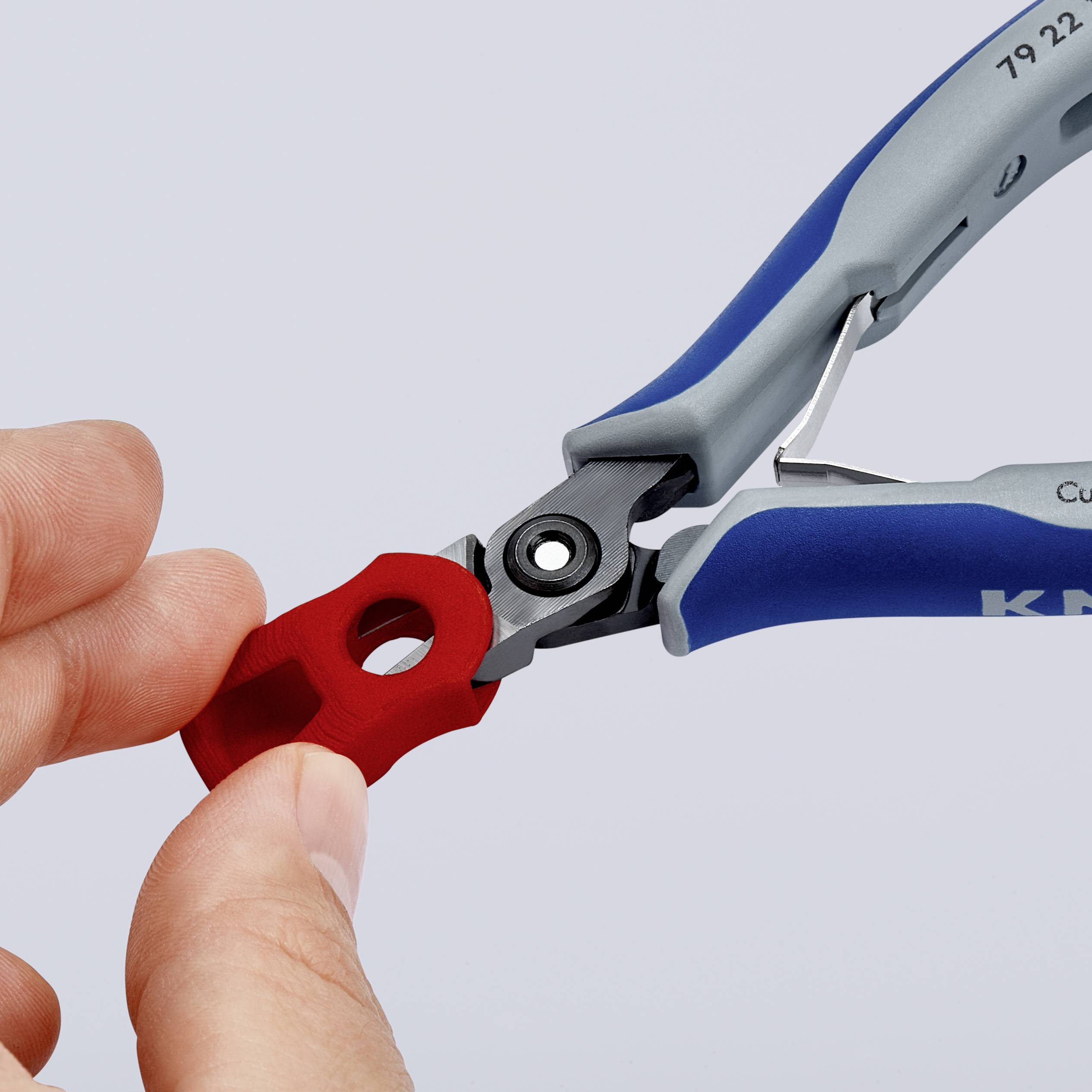 A hand holds a red wire cutter, while another hand holds a small red object near the blade.