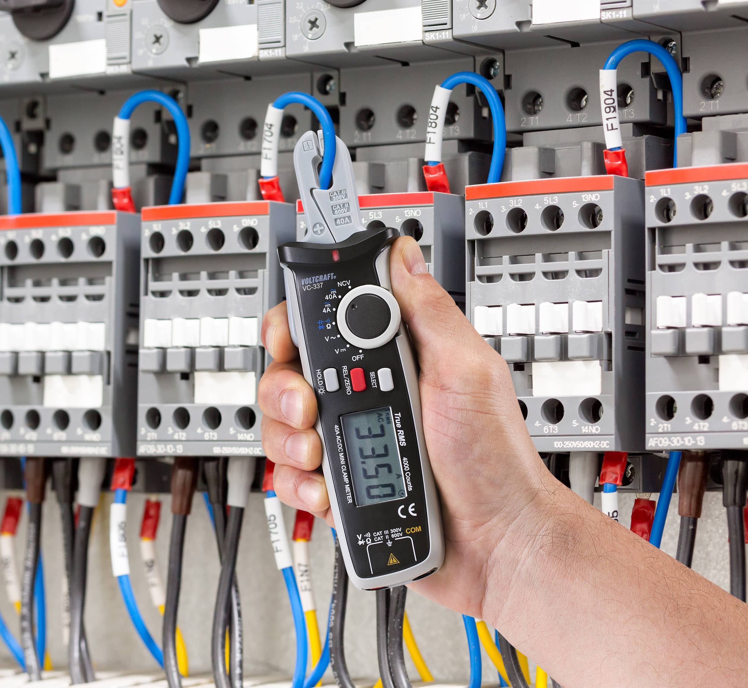 A hand holds a digital voltage meter in front of an open electrical panel. Colourful cables are connected to various switches.