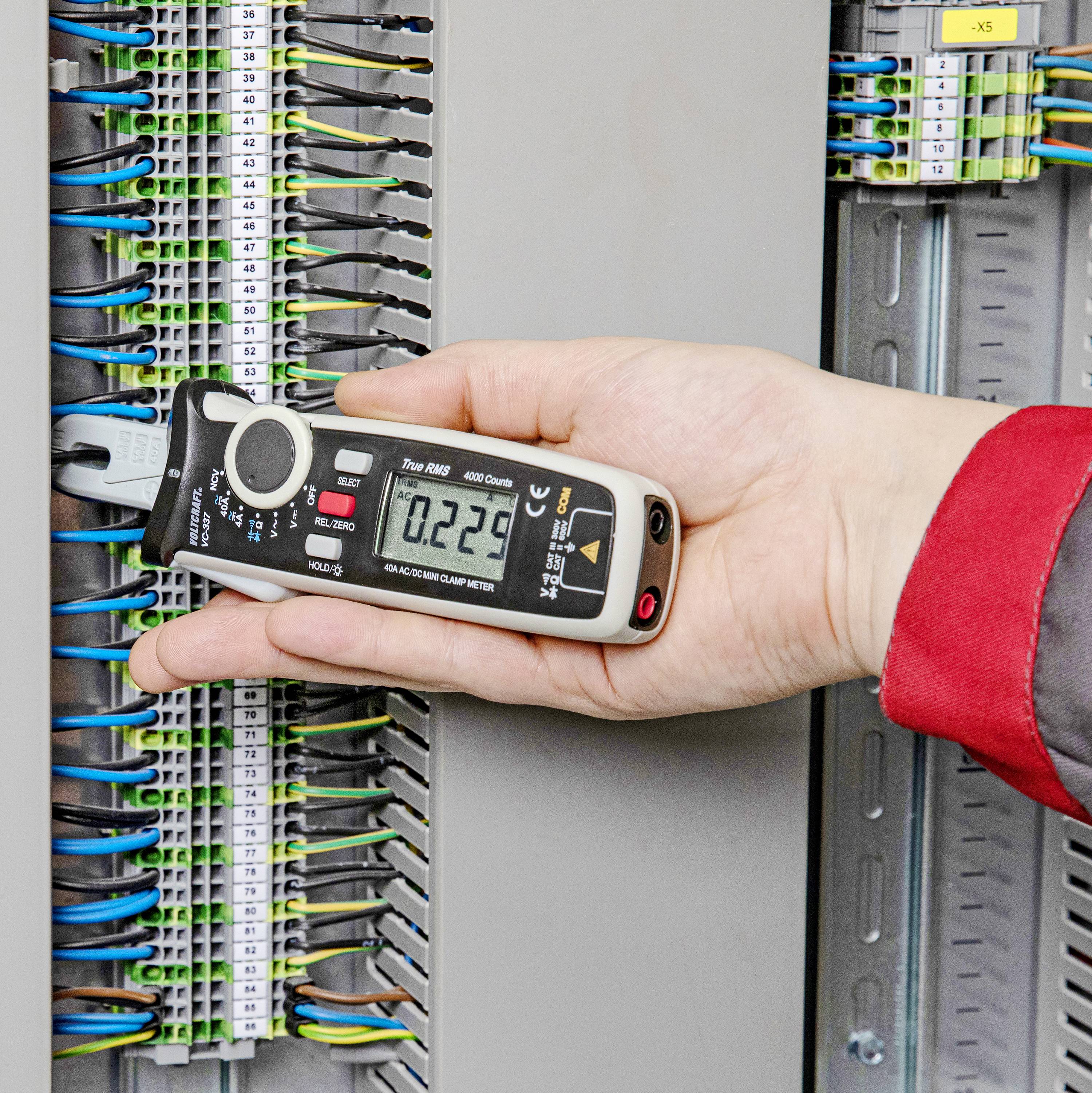 A person is holding a digital measuring device at an open electrical distribution board to measure electrical signals. Cables are visibly connected.