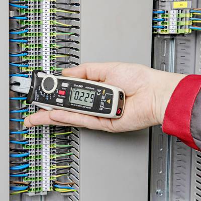 A person is holding a digital measuring device at an open electrical distribution board to measure electrical signals. Cables are visibly connected.