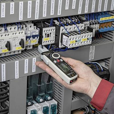 A hand is holding a digital multimeter and measuring the current in an open electrical cabinet with multiple wirings and devices.