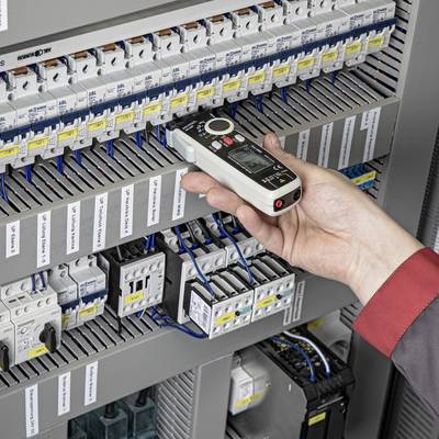 A hand is holding an electrical testing device and checking a fuse box with multiple switches and cables.