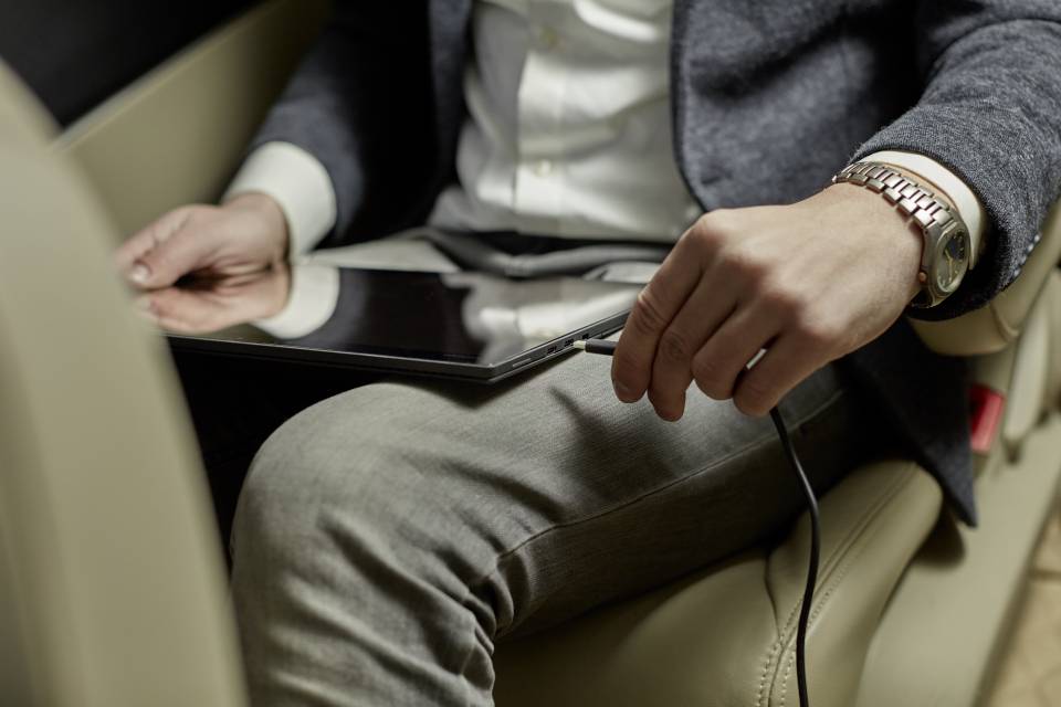 A person wearing a grey suit is holding a tablet connected by a cable while sitting in a car. The scene appears professional.