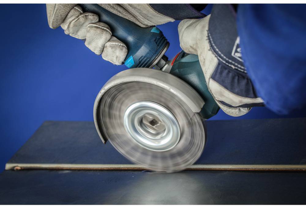 A person is wearing gloves and grinding a metal surface with an angle grinder. A blue wall is in the background.