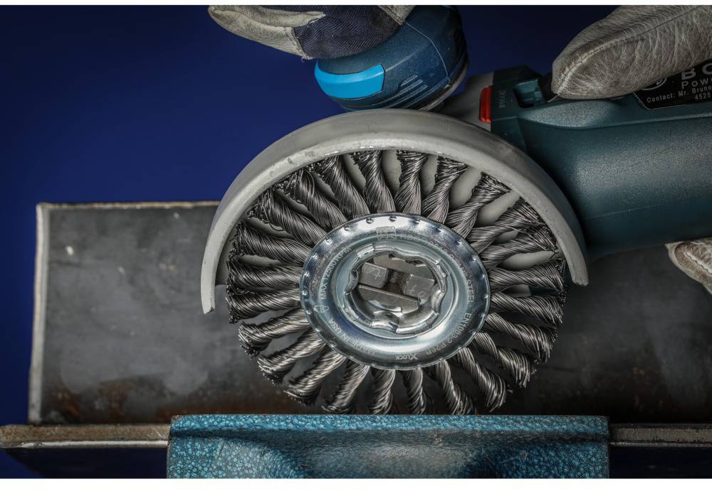 A wire brush on an angle grinder is cleaning the surface of a metal piece. A hand wearing work gloves is holding the tool.