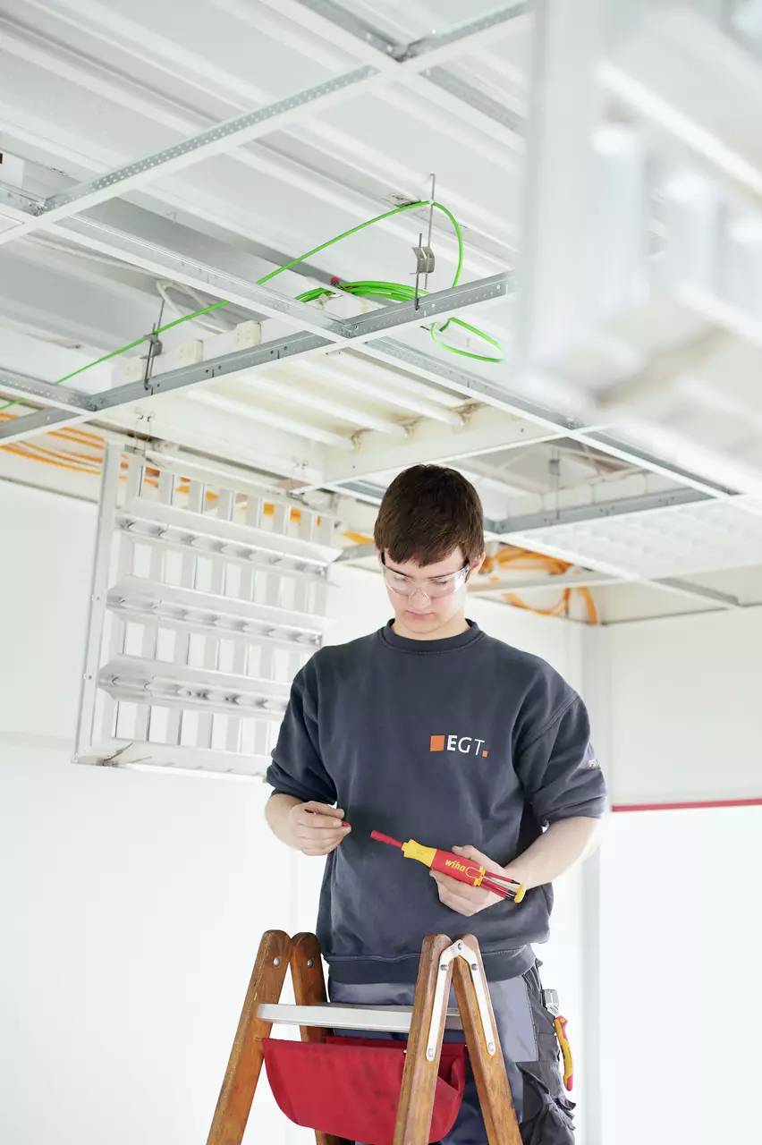 A person is standing on a ladder and working on an electrical ceiling. They are using tools and wearing work attire. Cables are visible.