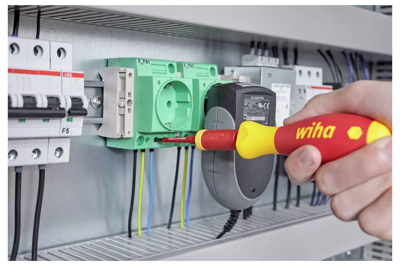 A hand uses a red and yellow Wiha screwdriver to tighten a green electrical outlet module inside an industrial control panel.
