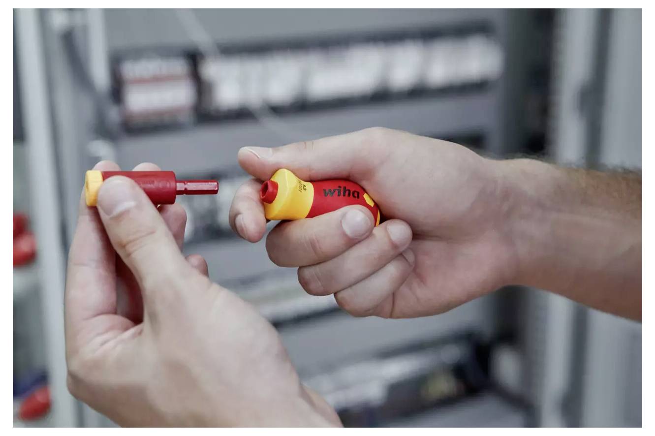 A person is holding a red and yellow screwdriver, assembling or disassembling its bits, with electrical equipment blurred in the background.