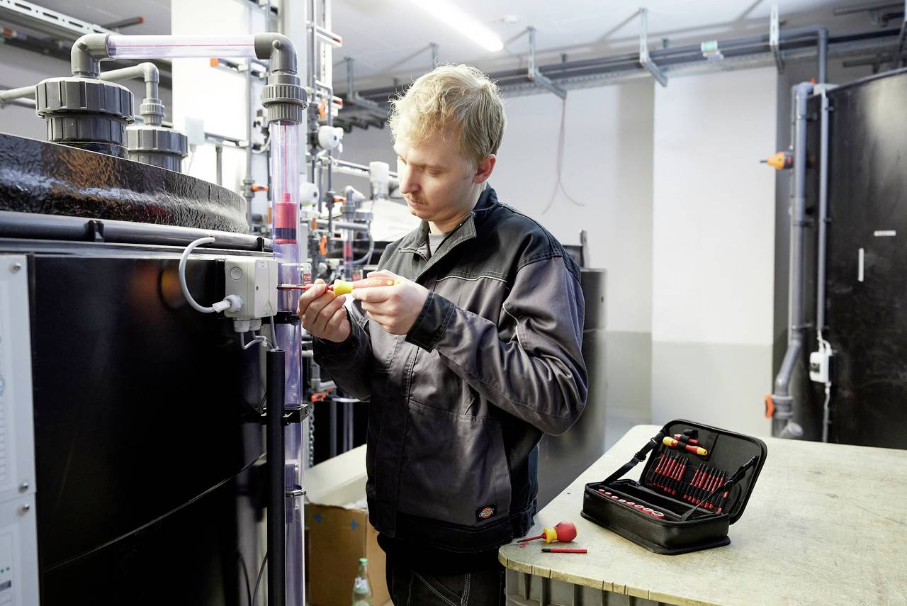 A technician in workwear is repairing equipment with a screwdriver. An open toolbox sits beside him on a table.