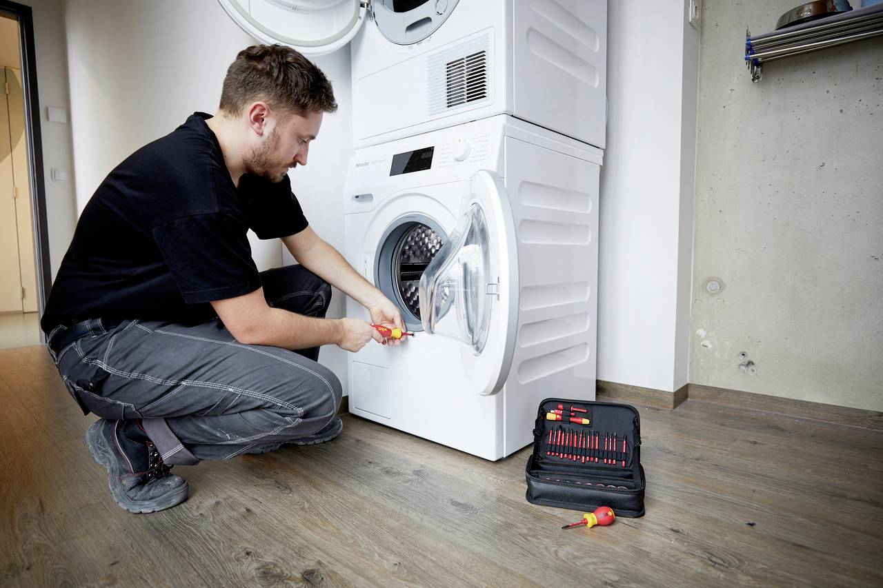 A man is repairing a washing machine with a screwdriver. A toolbox is lying on the floor. He is wearing work clothes and is focused on his task.