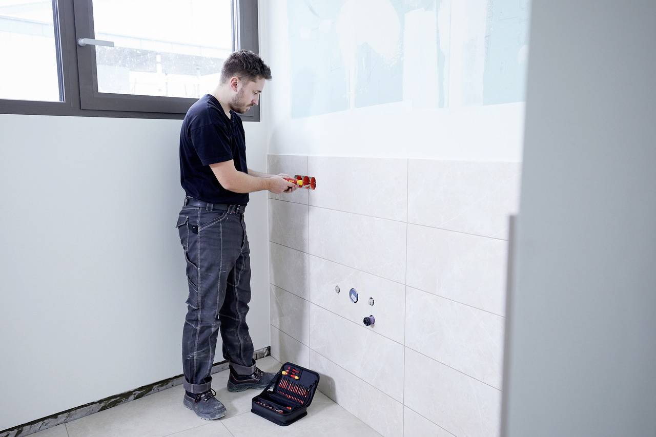 A man is working in a room with tiles, using a tool from a toolbox that is standing on the floor.