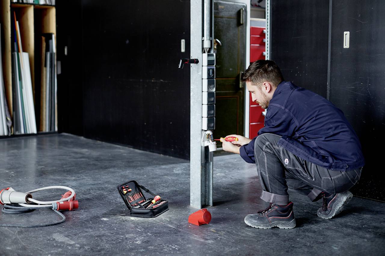 A man in workwear is repairing a metal door in a workshop. He is holding a tool in his hand. Shelves are visible in the background.