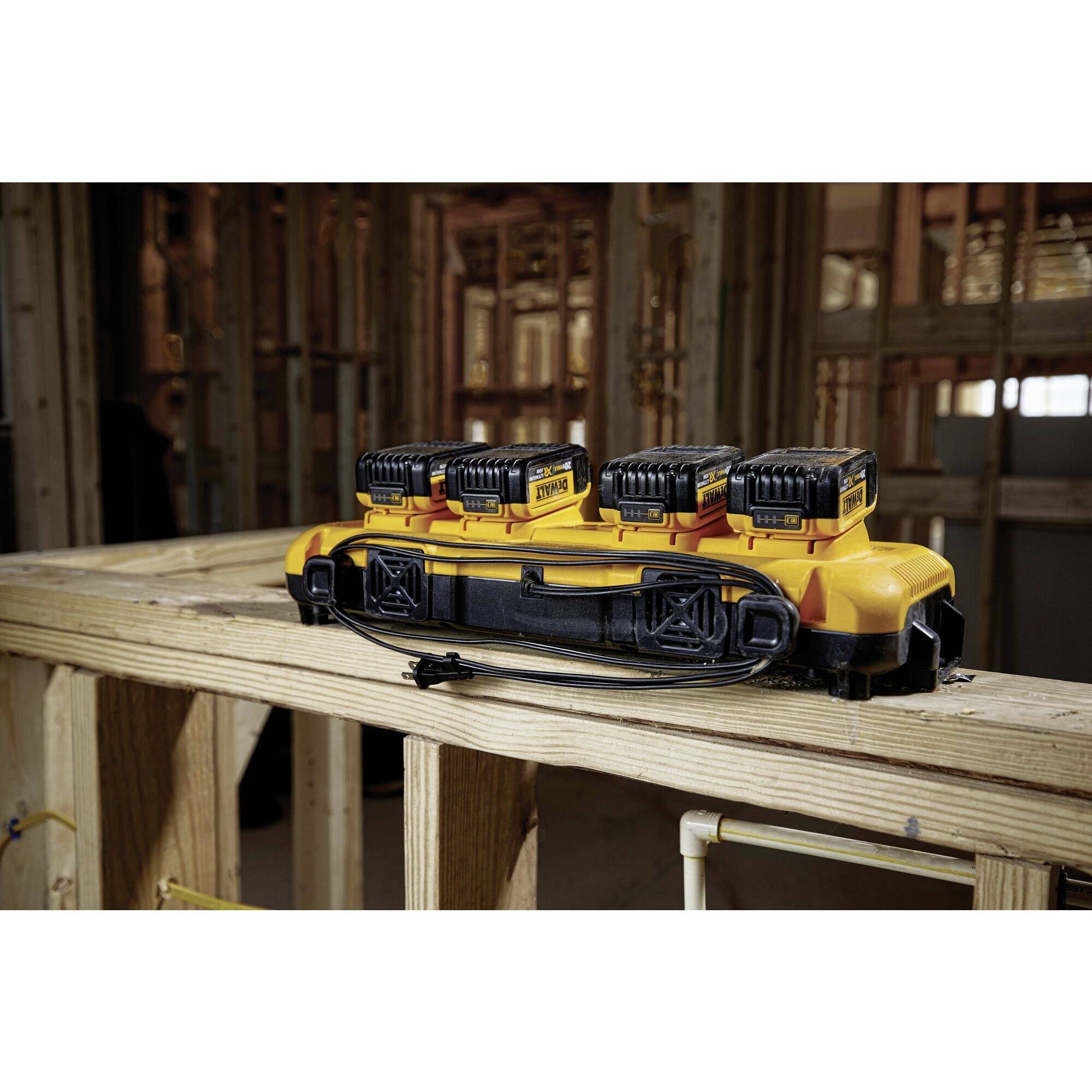 Four yellow batteries on a charger, placed on scaffolding in an unfinished room. Background shows wooden construction.