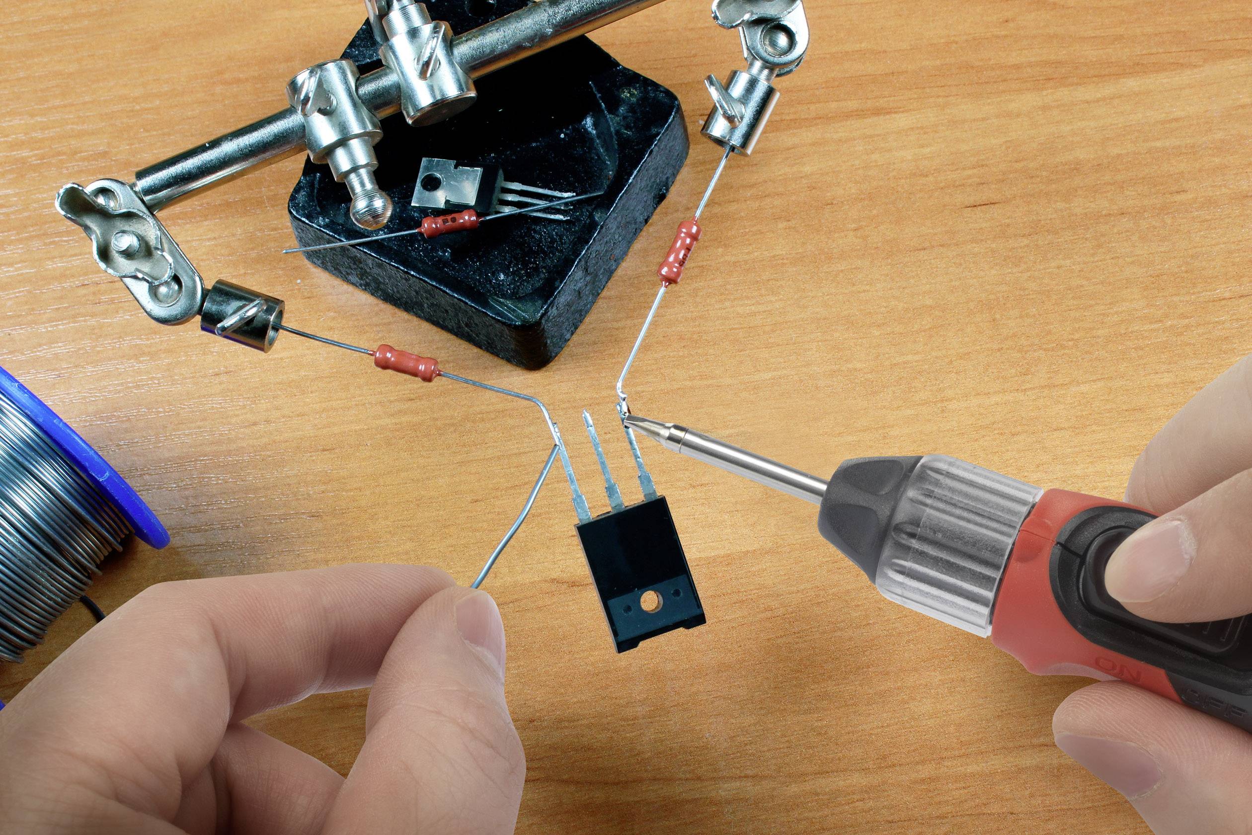 A person is soldering electronic components onto a circuit board. A soldering iron and several wires are visible.