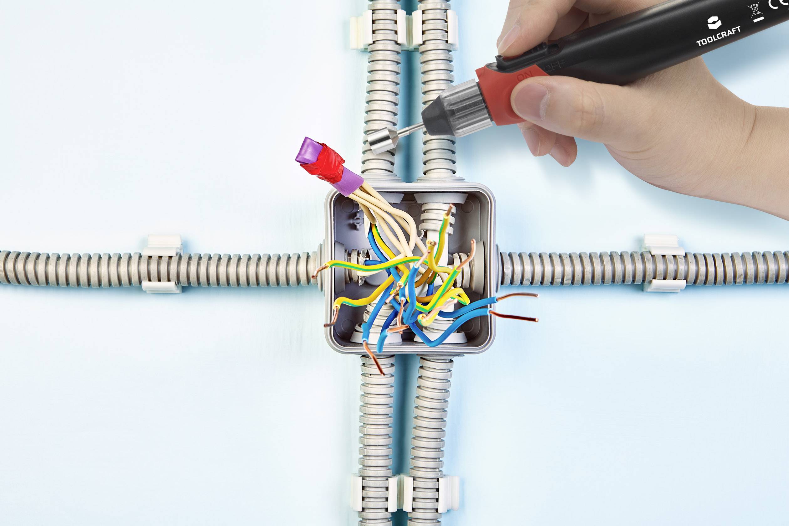 A hand holds a voltage tester over an open junction box with various coloured cables.